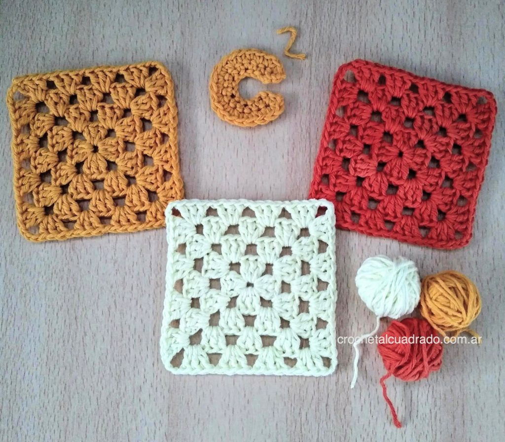 Tres cuadrados al crochet clásico en colores mostaza, amarillo y rojo sobre una mesa de madera