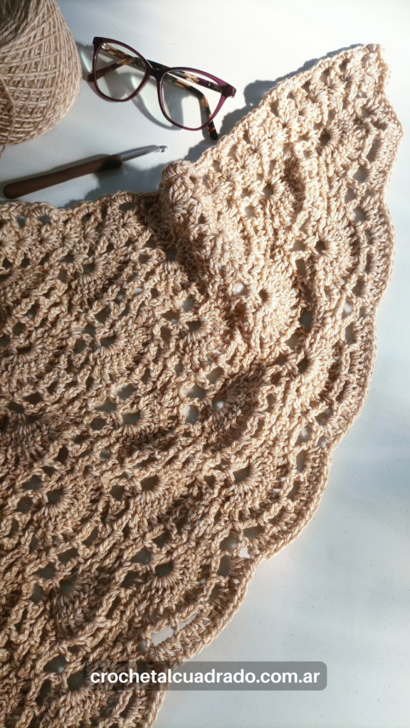 Chal triangular al crochet tejido con hilado grueso en color natural, con textura calada y gafas y ovillo sobre mesa blanca